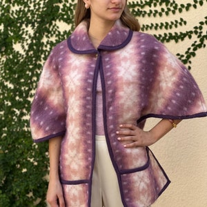 May include: A purple and white patterned cape with a rounded collar and pockets. The cape has a tie-dye effect with a purple trim. The model is wearing a striped shirt and white pants.