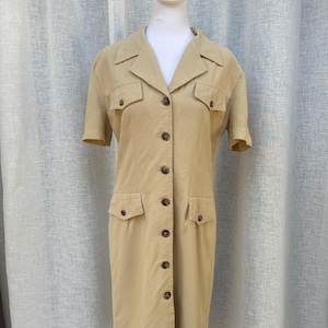 May include: A tan button-down dress with short sleeves, two chest pockets, and two hip pockets. The dress is displayed on a white mannequin and has a collared neckline. The dress is made of a light-colored fabric.