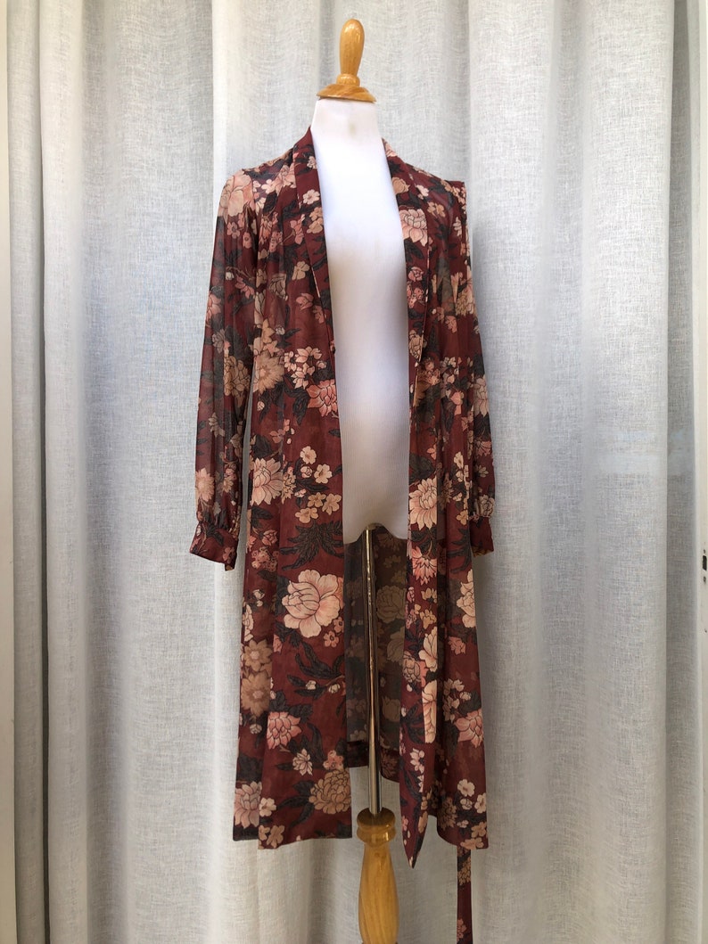 K&ouml;nnte beinhalten: Eine lange, maronfarbene, durchsichtige Robe mit Blumendruck. Die Robe hat einen Schalkragen und einen Bindeg&uuml;rtel.