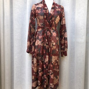 K&ouml;nnte beinhalten: Ein lang&auml;rmeliges, blumiges Kleid mit V-Ausschnitt und Bindeg&uuml;rtel. Das Kleid ist aus einem durchsichtigen Stoff gefertigt und hat eine lockere Passform.