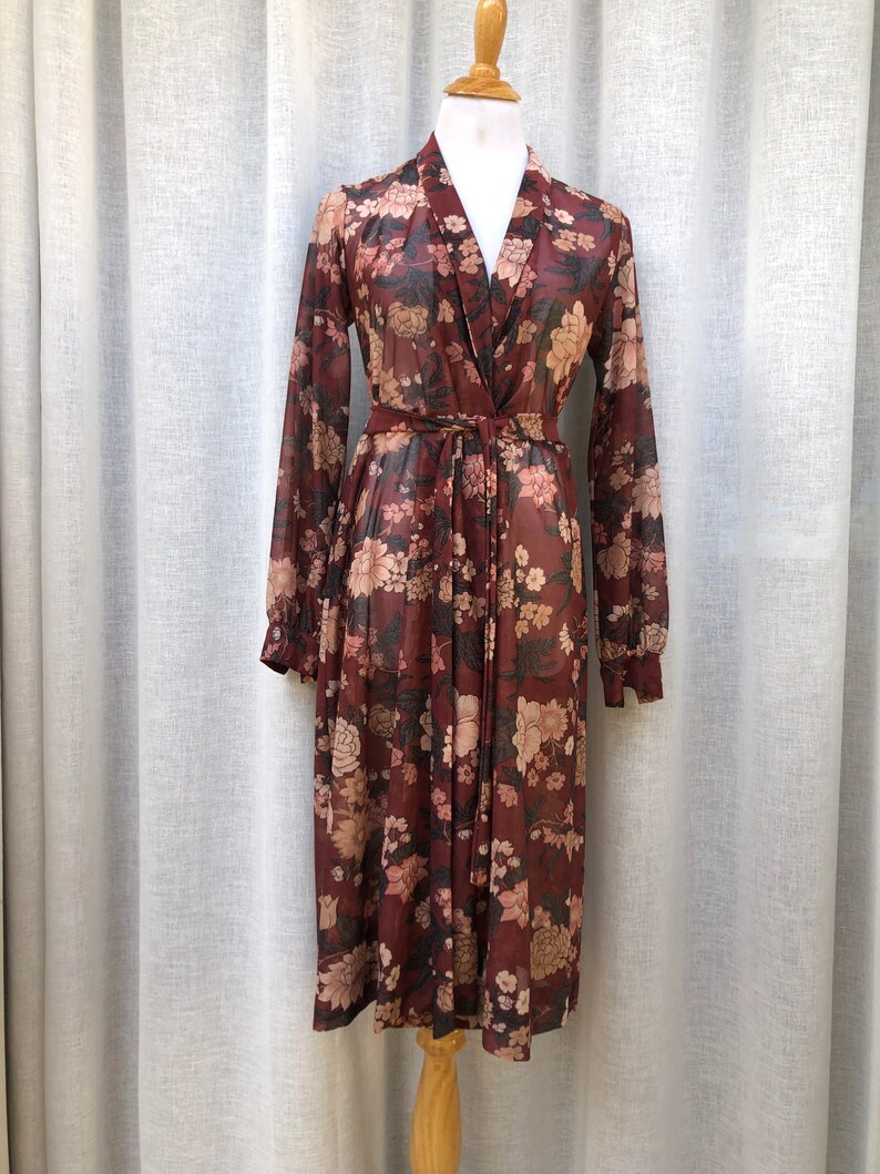 K&ouml;nnte beinhalten: Ein lang&auml;rmeliges, blumiges Kleid mit V-Ausschnitt und Bindeg&uuml;rtel. Das Kleid ist aus einem durchsichtigen Stoff gefertigt und hat eine lockere Passform.