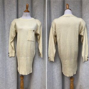 May include: A cream-colored knitted sweater with long sleeves and a patch pocket on the front. The sweater has gold buttons on the cuffs.