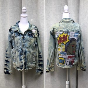 May include: A light blue acid-wash denim jacket featuring a distressed collar and a large, colorful pop-art graphic on the back. The graphic depicts a stylized person with the text WOW and OOPS printed in a comic book style on a white background.