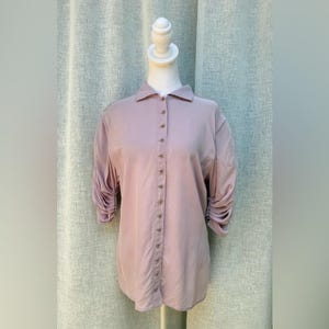 May include: A light mauve button-down shirt with three-quarter sleeves. The sleeves are gathered at the elbow. The shirt has a classic collar and is buttoned down the front with small, round buttons. The shirt is displayed on a mannequin.