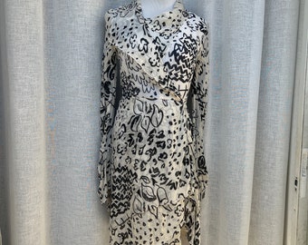 May include: A white dress with a black and white abstract print. The dress has a long sleeve and a wrap style. The dress is on a mannequin.