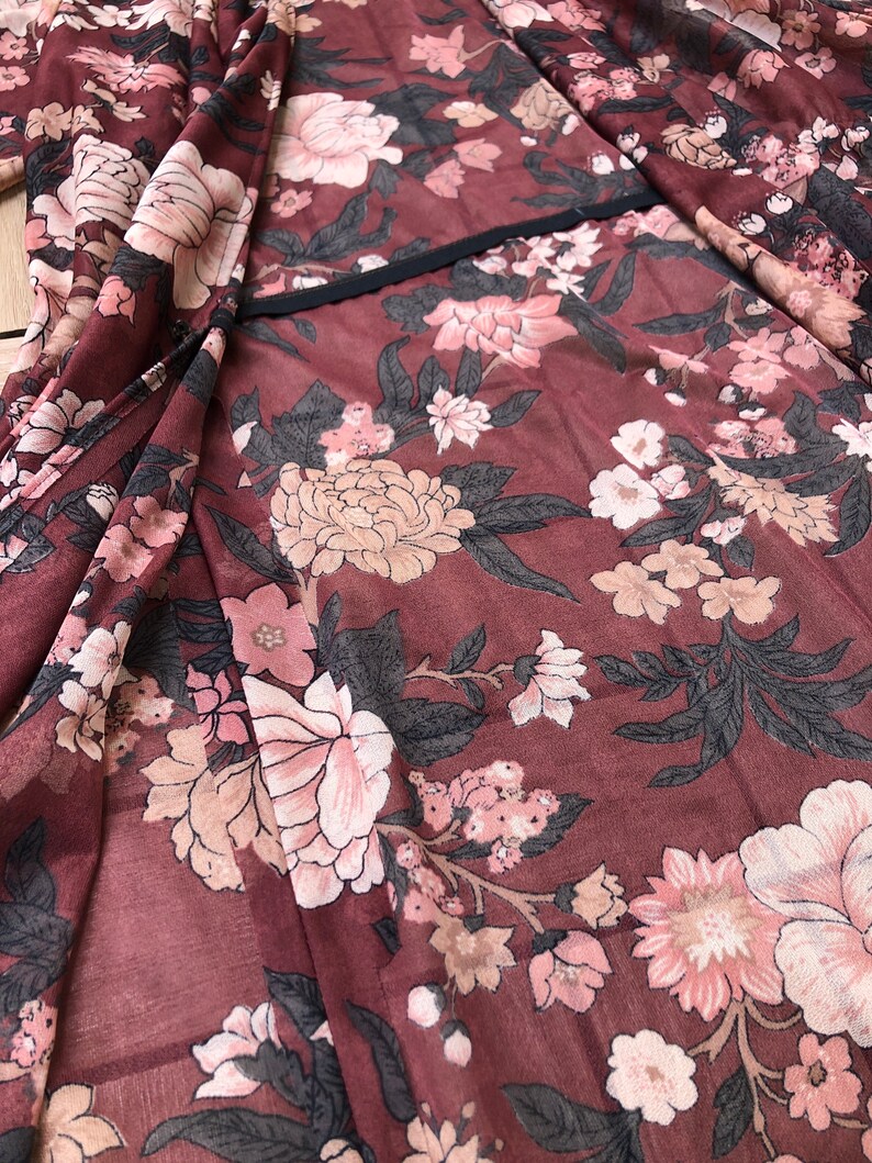 K&ouml;nnte beinhalten: Ein maroner, durchsichtiger Stoff mit einem floralen Muster in Rosa- und Beiget&ouml;nen. Der Stoff hat einen schwarzen Saum entlang der Kante.