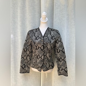 May include: A blazer with a snakeskin pattern in shades of gray and black. The jacket has a V-neckline, long sleeves, and is buttoned down the front. The edges are trimmed with black piping.