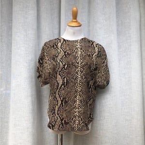 May include: A short-sleeved top with a snakeskin pattern in shades of brown and tan. The blouse is displayed on a mannequin. The fabric has a textured appearance, and the design is a repeating pattern.
