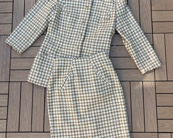 May include: A two-piece houndstooth suit in tan and white. The jacket has a round collar and button closure. The skirt is knee-length with a straight cut and two front pockets. The suit is made of a woven fabric.