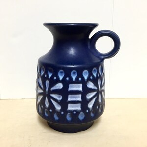 May include: A blue ceramic vase with a white floral design and a handle. The vase has a textured surface and a rounded shape.