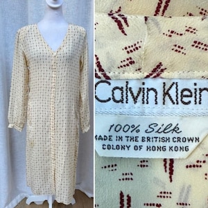 May include: Cream-colored Calvin Klein silk dress with a V-neck and long sleeves. The dress features a button-down front and a subtle pattern of small, dark red shapes. The label reads "100% Silk" and "MADE IN THE BRITISH CROWN COLONY OF HONG KONG."
