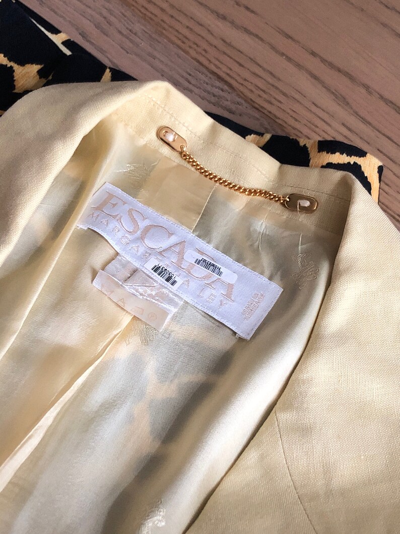 May include: A close-up of the inside of a yellow blazer with a black and gold patterned lining. The label reads "ESCA" and "MADE IN ITALY".