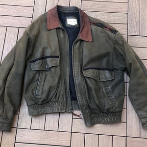 May include: Olive green leather bomber jacket with a brown collar and black accents. Features two front pockets and decorative stitching. The jacket has a ribbed hem and cuffs, and a zipper closure.