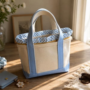 May include: A small canvas tote bag with light blue accents. The bag has a cream-colored body, light blue handles and trim, and a blue and white gingham lining. Other items include a blue scrunchie, a small blue wallet, and sunglasses.
