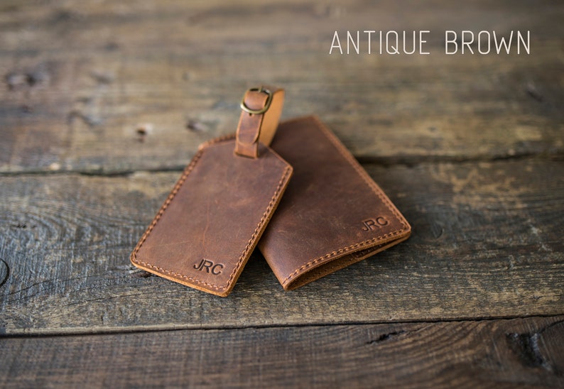 May include: Two brown leather luggage tags with the initials "JRC" embossed on them. The tag on the left has a brass loop for attaching to luggage. The text "ANTIQUE BROWN" is visible at the top of the image.