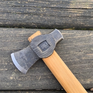 May include: A black and silver hand axe with a wooden handle. The axe head has a small, rounded blade and a hammer head on the opposite side. The axe head is stamped with the letters "B.I.D."