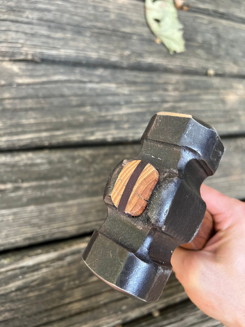 May include: A black metal hammer with a wooden handle. The hammer head has a flat striking surface and a rounded face. The handle is wrapped in brown leather.