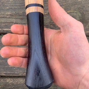 May include: Close-up of a wooden axe handle with a black stain and two black bands. The handle is held in a hand.