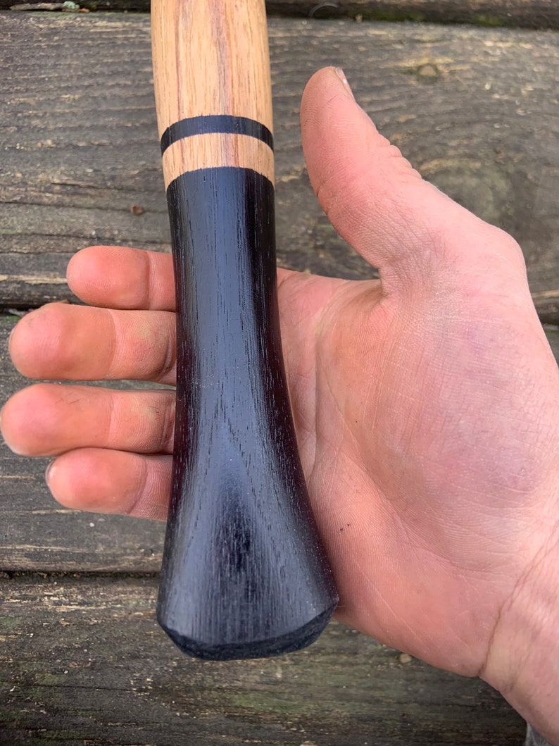 May include: Close-up of a wooden axe handle with a black stain and two black bands. The handle is held in a hand.