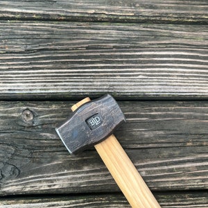 May include: A black metal hammer with a wooden handle. The hammer head has a square shape with the letters "BLD" stamped on it.