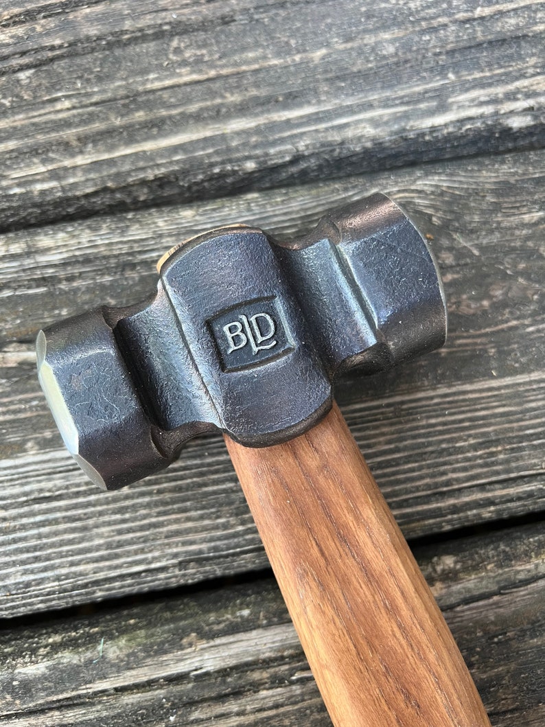 May include: A black metal hammer with a wooden handle. The hammer head has a rounded face and a smaller, square face. The hammer is marked with the letters "BLD".