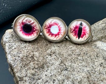 May include: Three silver rings featuring circular cabochons with realistic eye designs. Each cabochon displays a vibrant pink and white iris pattern with distinct pupil shapes, including a round black pupil, a white center, and a vertical slit pupil.