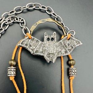 May include: A silver-toned bat pendant necklace with an orange cord and decorative beads. The bat pendant has a textured surface and is suspended from a silver chain. A brass-colored arch is above the bat.
