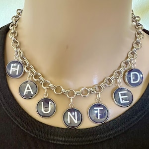May include: Silver-toned chain necklace with circular pendants spelling "HAUNTED". Each pendant has a dark blue background with a white letter. The necklace is displayed on a mannequin.
