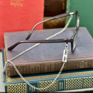 May include: Dark-framed eyeglasses with a silver chain, resting on a stack of vintage books. The chain features a rectangular charm with an eye design. The books are various colors, including red, blue, and green.