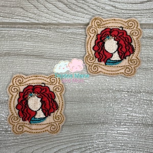 May include: Two embroidered patches featuring a cartoon woman with red curly hair in a gold picture frame. The patches are on a light brown wood background.