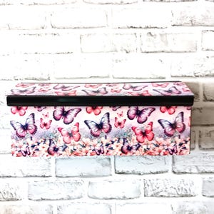 May include: Rectangular storage box with a black lid and a vibrant butterfly pattern. The box features pink, purple, and white butterflies against a floral background. The box is mounted on a white brick wall.