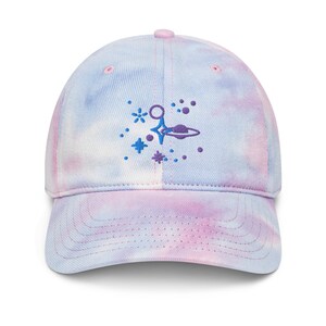 Puede incluir: Gorra de béisbol teñida con tinte anudado en tonos azul claro, rosa y morado. La gorra presenta un diseño bordado de un planeta, estrellas y otros elementos celestes en azul y morado.