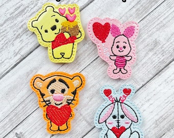 May include: Four embroidered felt patches featuring Winnie the Pooh, Piglet, Tigger, and a bunny rabbit. Each character is holding a red heart. The patches are on a light wood background.