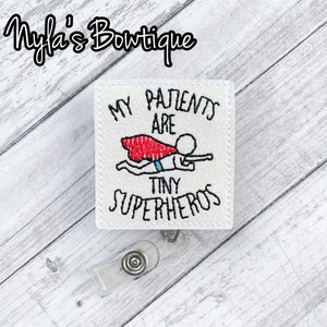 May include: White embroidered badge with the text "My patients are tiny superheroes" and a cartoon superhero flying with a red cape.