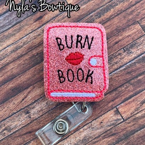 May include: A pink glitter badge reel with the words "BURN BOOK" in black, a red lip graphic, and a clear plastic clip. The badge reel is rectangular and has a stitched border. The text "Nyla's Bowtique" is in the upper left corner.