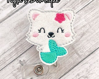 May include: White embroidered polar bear with a pink flower on its head and a teal mermaid tail. The bear is holding the tail in its paws. The bear is attached to a clear plastic badge holder.