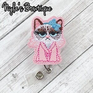 May include: Pink embroidered cat with a blue bow, wearing a pink robe with a white letter M, attached to a retractable badge holder.