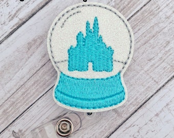 May include: A white glitter snow globe with a blue embroidered castle inside. The snow globe is attached to a clear plastic badge holder.