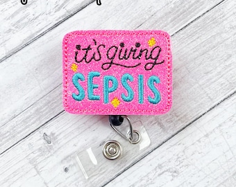 May include: A pink badge reel with the phrase "it's giving SEPSIS" embroidered in black and teal thread. The badge reel has a clear plastic holder and a metal clip. The top left corner of the image has the text "Nyla's Boutique".