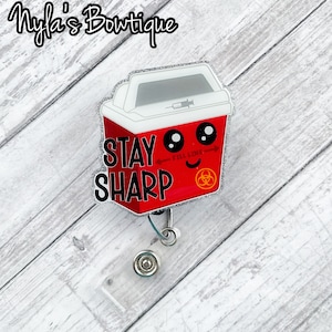 May include: Red and white cartoon sharps container with a biohazard symbol and the text "Stay Sharp" and "Fill Line". The container has googly eyes and a smiling mouth. The container is attached to a silver retractable badge reel.