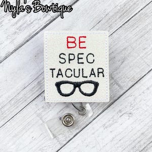May include: A white badge reel with the words "BE SPECTACULAR" in black and red, and a pair of black glasses. The badge reel has a clear plastic strap and a metal clip. The top left corner has the text "Nyla's Boutique".