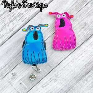 May include: Two colorful ghost-shaped retractable badge holders. The blue ghost has a black face and orange antennae. The pink ghost has a black face and orange antennae. Both ghosts are on a wood background.