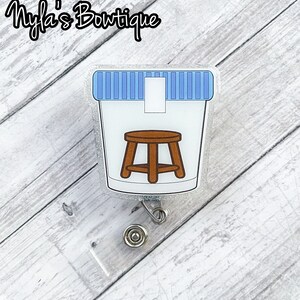 May include: A silver glitter badge reel with a clear plastic charm featuring a white cup with a blue lid and a brown stool inside. The charm is attached to a clear plastic badge holder.