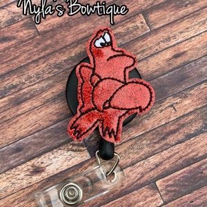 May include: Red glitter Sebastian the crab from The Little Mermaid character on a black retractable badge reel with a clear plastic badge holder.