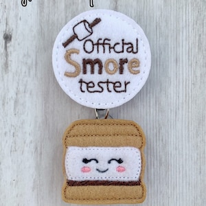 May include: White felt circle with brown embroidery that reads "Official S'more Tester" with a stick figure holding a marshmallow. A brown felt s'more with a white marshmallow and brown chocolate is attached to the circle.