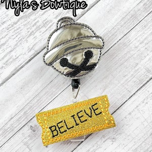 May include: A silver jingle bell with a black outline and a gold ticket with the word "BELIEVE" on it. The bell and ticket are attached to a retractable badge holder.