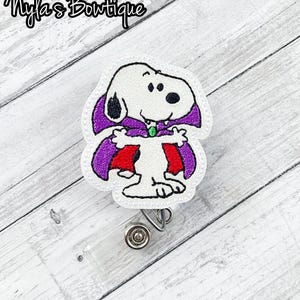 May include: A white Snoopy badge reel with a purple cape and red lining, designed as a vampire. The badge reel has a clear plastic holder and a metal clip. The text "Nyla's Boutique" is in the upper left corner.