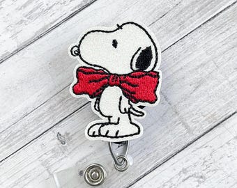 Könnte beinhalten: Ein weißes Snoopy-Ausweis-Jojo mit roter Fliege. Das Ausweis-Jojo hat eine transparente Kunststoffbasis und einen Metallclip. Der Text "Nyla's Boutique" befindet sich in der oberen linken Ecke.