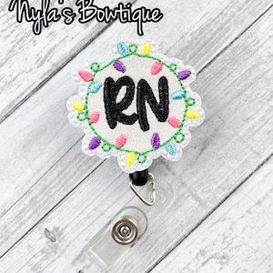 May include: A white embroidered badge with a circle of colorful lights around the letters "RN". The badge is attached to a black retractable badge holder.