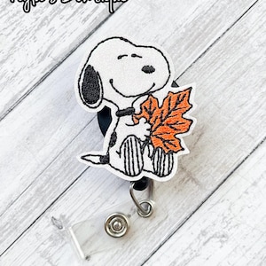 May include: A badge reel featuring an embroidered Snoopy character holding a large orange autumn leaf. The design is outlined in black thread on a white background. The text Nyla's Bowtique is visible in the upper left corner. The reel has a clear plastic strap with a metal snap button.